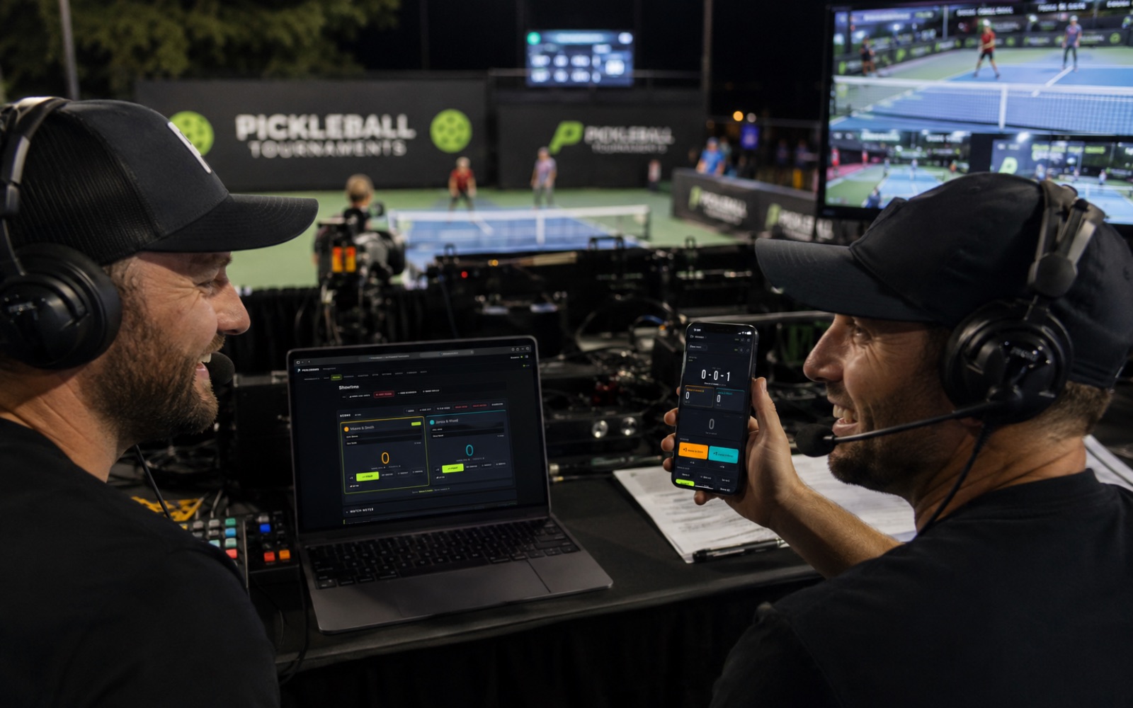 Commentators calling a match from the PickleBoard booth — the control panel on a laptop, live scoring on a phone, the match on the big screen